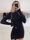 Woman wearing a black turtleneck bodycon mini dress with long sleeves and ruched details, posing in a boutique.